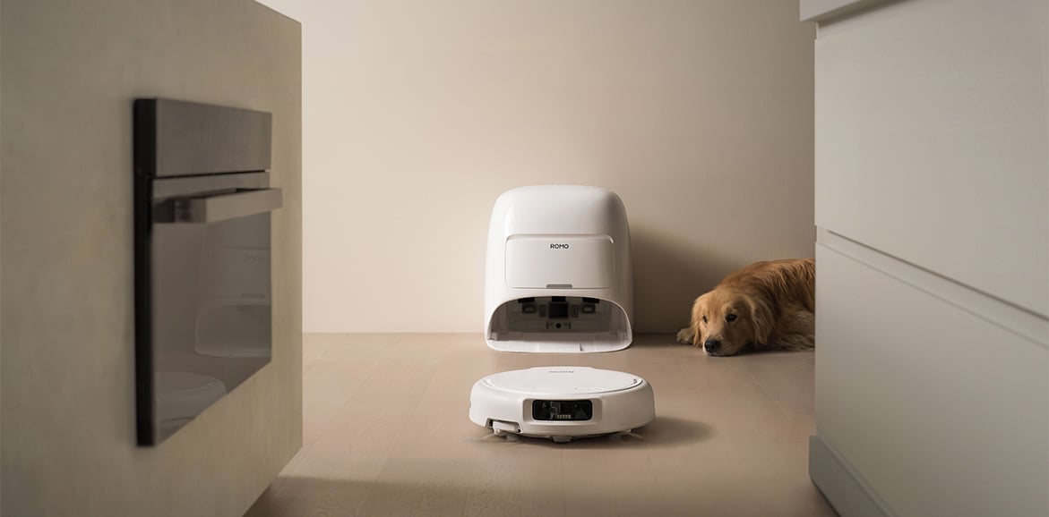 Robot sprzątający DJI ROMO S wyjeżdża ze stacji dokujacej między meble a obok leży pies